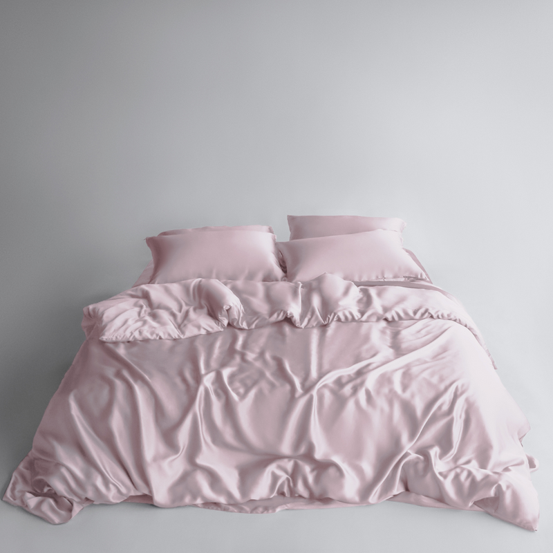 A neatly made bed features the Bedding Bundle in light pink silky fabric with two matching pillows, set against a plain white background. The slightly rumpled blanket adds a soft and inviting look.