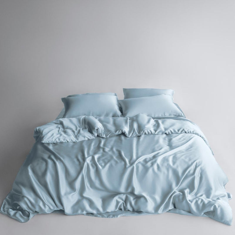 A neatly made bed with the Bedding Bundle - light blue silky cooling bedding, two pillows, and matching sheets - set against a plain light grey background.