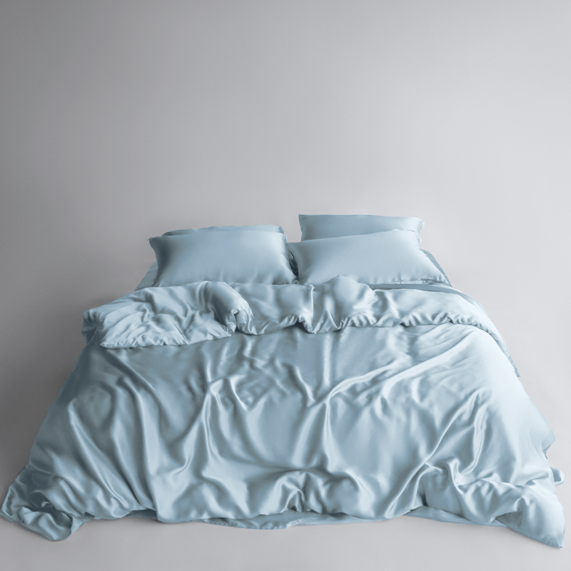A neatly made bed with the Bedding Bundle - light blue silky cooling bedding, two pillows, and matching sheets - set against a plain light grey background.