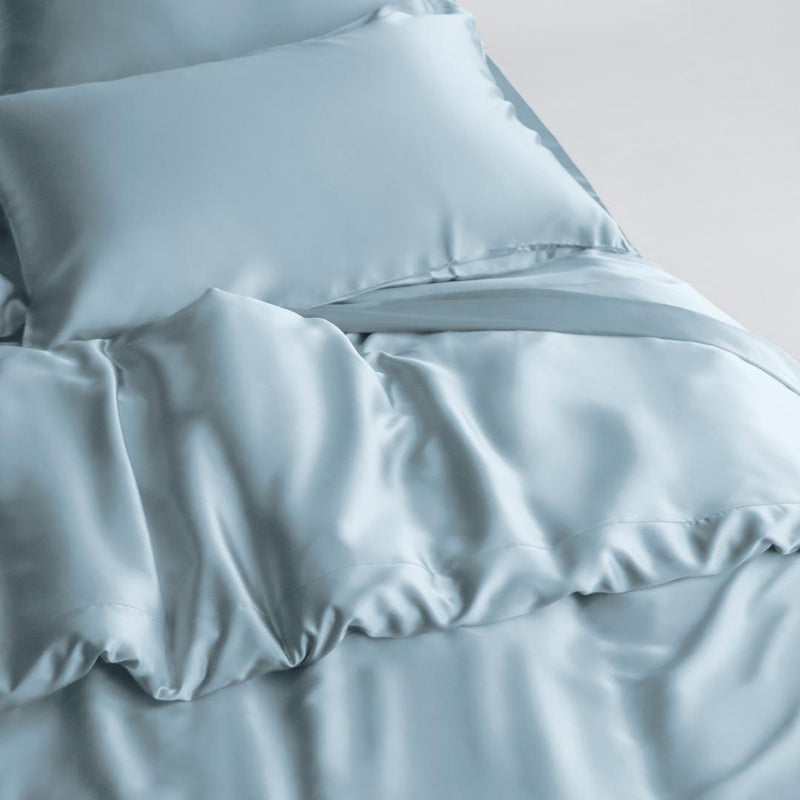 A bed is neatly made with a light blue silky Duvet Cover, featuring a matching pillow and sheets all showcasing a smooth, shiny fabric texture.