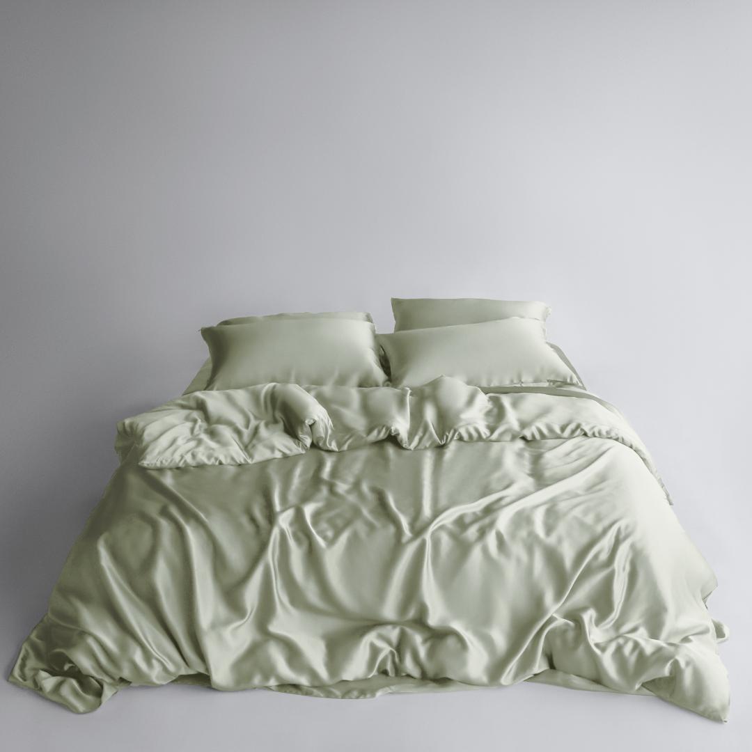 A neatly made bed features the Bedding Bundle in sage green: silky, cooling bedding with two matching pillows. The smooth fabric has natural folds and a soft texture, all set against a plain white background.