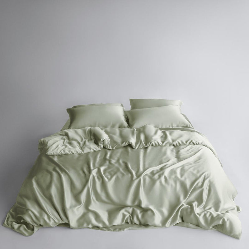 A neatly made bed features the Bedding Bundle in sage green: silky, cooling bedding with two matching pillows. The smooth fabric has natural folds and a soft texture, all set against a plain white background.