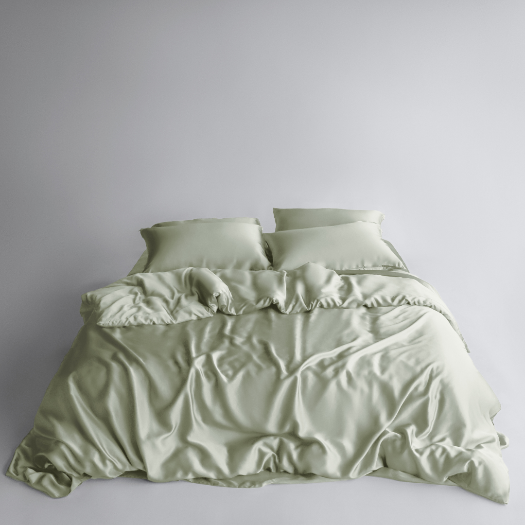 A neatly made bed features the Bedding Bundle in sage green: silky, cooling bedding with two matching pillows. The smooth fabric has natural folds and a soft texture, all set against a plain white background.