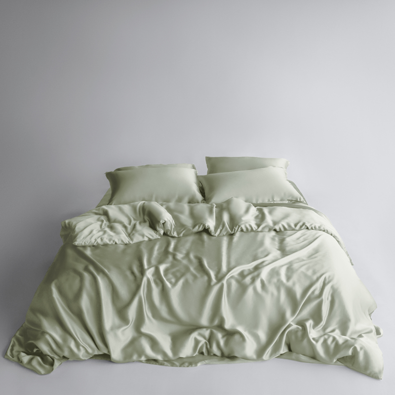 A neatly made bed features the Bedding Bundle in sage green: silky, cooling bedding with two matching pillows. The smooth fabric has natural folds and a soft texture, all set against a plain white background.