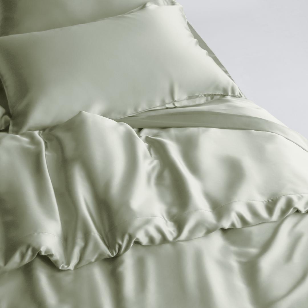 A neatly made bed features a Duvet Cover in soft, sage green silky fabric with matching pillowcases. The smooth, shiny fabric and gentle folds create a luxurious look, while sustainable bedding enhances comfort and style for an inviting space.