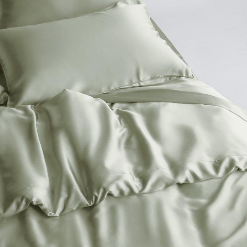 A neatly made bed features a Duvet Cover in soft, sage green silky fabric with matching pillowcases. The smooth, shiny fabric and gentle folds create a luxurious look, while sustainable bedding enhances comfort and style for an inviting space.