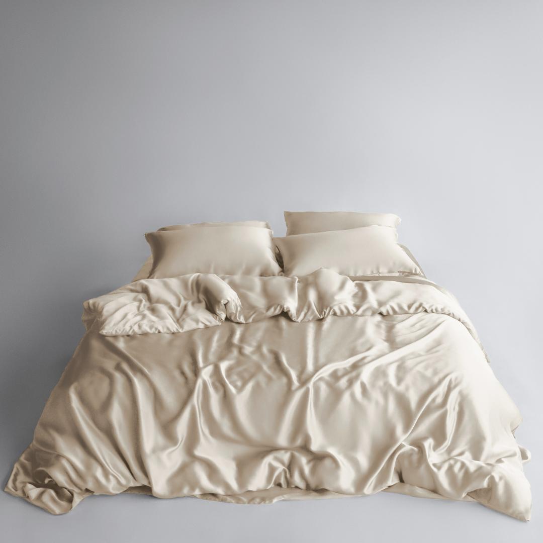 A neatly made bed features a sustainable Duvet Cover, sand-coloured sheets, and two pillows, all set against a plain light grey background.