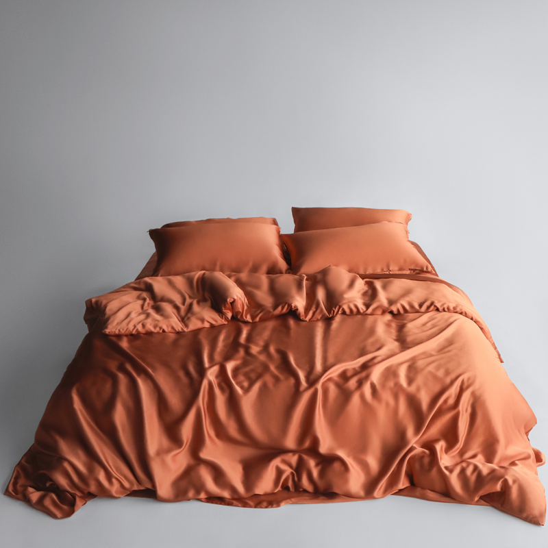 A neatly made bed with terracotta silky sheets, duvet cover and two pillows, set against a plain light grey background.