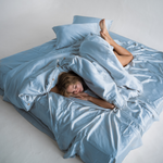 A person lies comfortably on a large bed covered in light blue, shiny sheets and pillows, partially wrapped in the bedding, in a bright, minimalistic room.