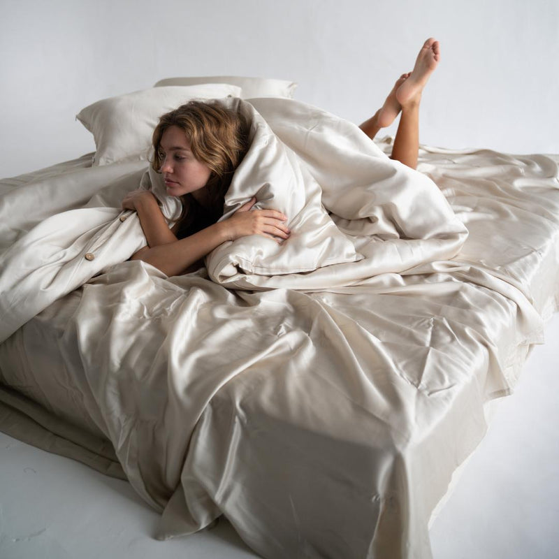 A young woman relaxes on a softly lit bed covered in a luxurious sand-coloured Bedding Bundle, with her legs raised and crossed, looking cozy in a minimal setting.
