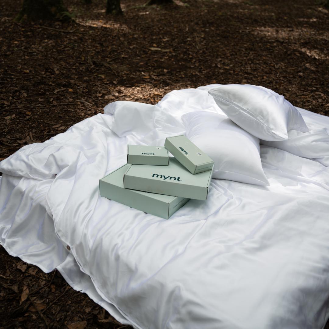 A white silky bedspread and pillow rest on a forest floor, with several light green boxes labeled mynt lab - each containing the Bedding Bundle for cooling comfort - arranged on the bedspread under natural daylight.