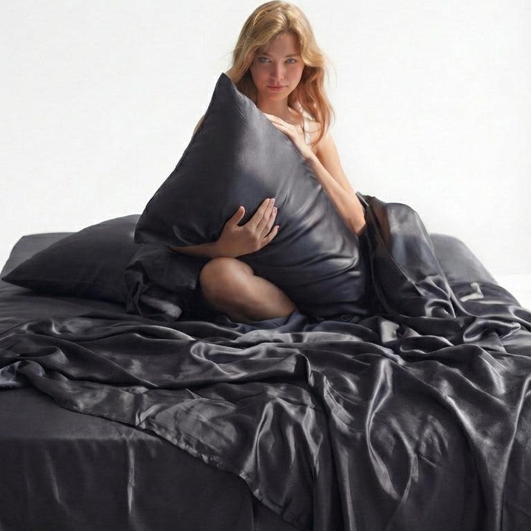 A woman sits on a bed covered in Mynt Lab’s MyntFusion™ Complete Sheet Set (4-Piece), holding a matching pillow. The silky, cooling bedding is smooth and shiny - ideal for hot sleepers - in a softly lit, minimalistic setting.
