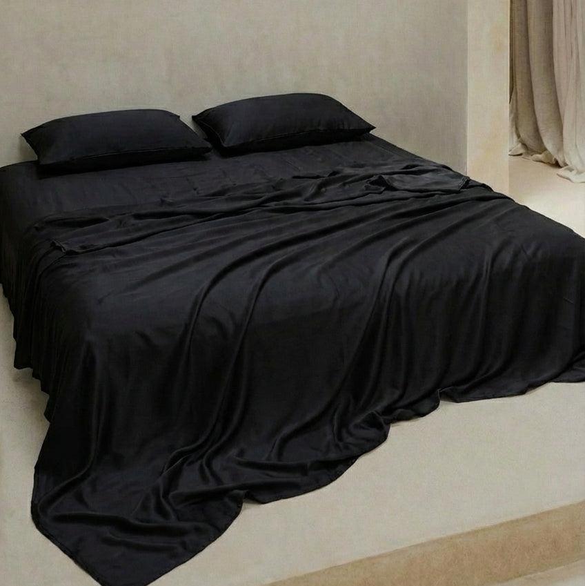 Experience perfect comfort for hot sleepers with the Mynt Lab MyntFusion™ Complete Sheet Set (4-Piece) in black  - includes two pillows, cooling sheets, and a flowing blanket, all set against a plain beige wall.