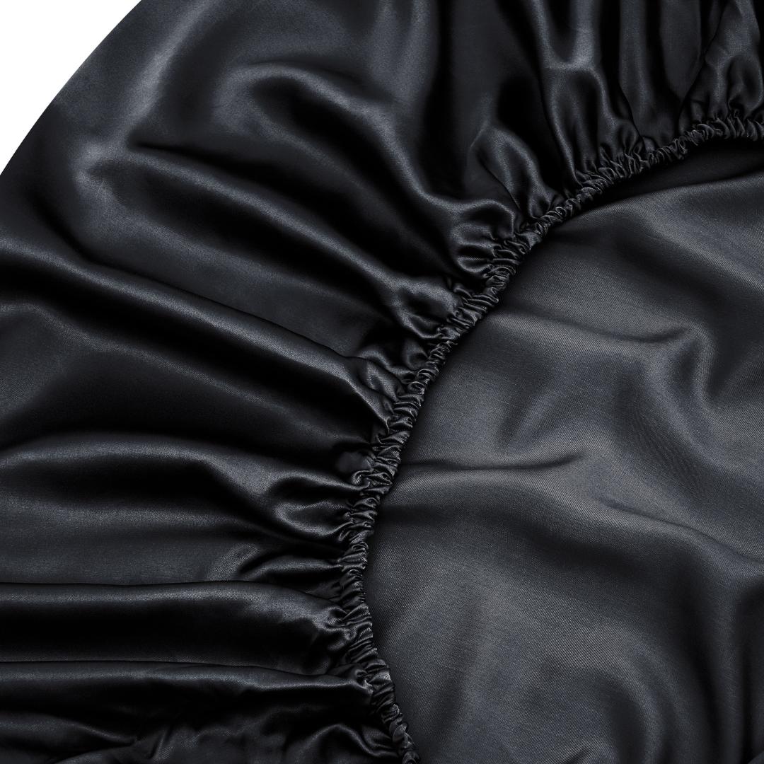 Close-up of Mynt Lab's Black Fitted Sheet Set (Super King) shows smooth, shiny satin fabric with gathered elastic edges, offering a deep pocket fit and a cooler feel compared to cotton.