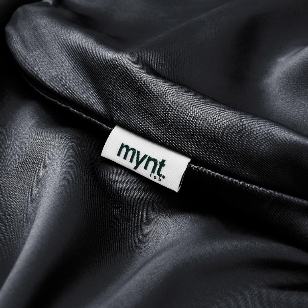 A close-up of the Mynt Lab Black Fitted Sheet Set (Super King) reveals its lustrous satin fabric with a white "mynt. lab." tag, offering a soft, cool surface that regulates temperature for comfort superior to cotton.
