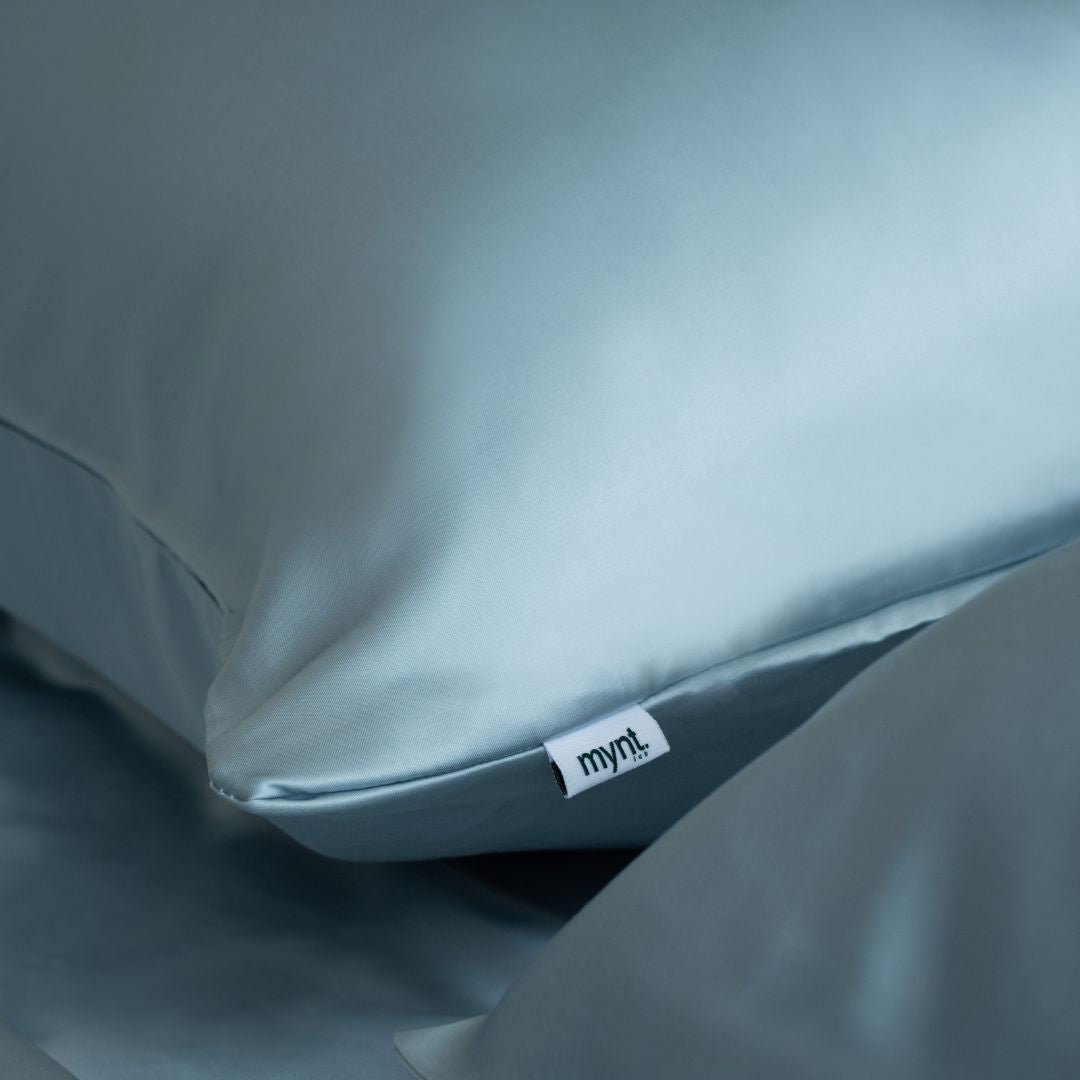 Close-up of light blue vegan silk pillowcase with visible Mynt Lab fabric tag, styled on matching silky bedding