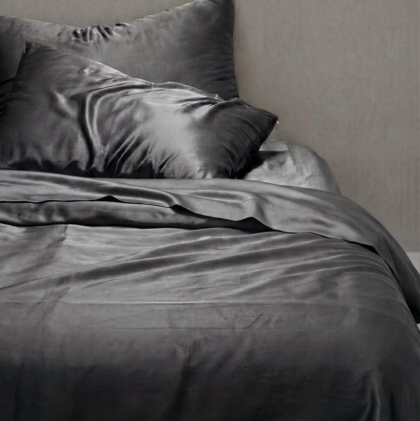 A bed dressed in Mynt Lab's MyntFusion™ Complete Sheet Set (4-Piece) in smooth, shiny dark grey with matching pillowcases sits against a light grey wall. This hypoallergenic bedding is soft, cool, and ideal for hot sleepers.