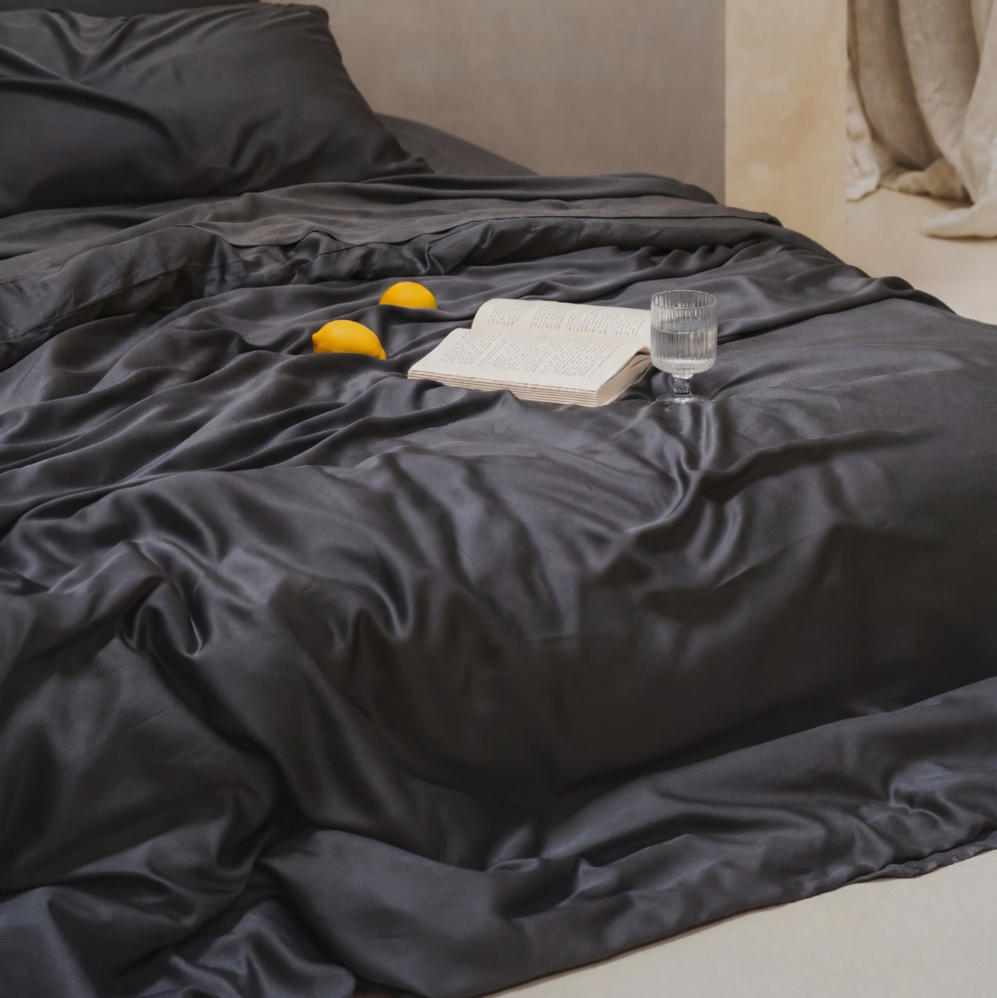 A Mynt Lab MyntFusion™ Duvet Cover rests on a bed with black silky sheets, an open book, a glass of water, and three lemons - perfect for hot sleepers in a soft, rumpled look within a minimalist, neutral-toned room.