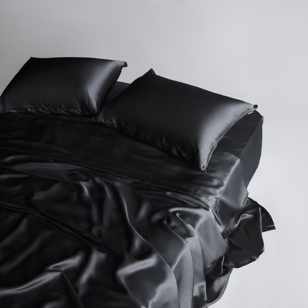 The Complete Sheet Set (4-Piece) features silky soft black sheets and two matching pillows, styled on a neatly made bed against a plain white background for a sleek, modern look.