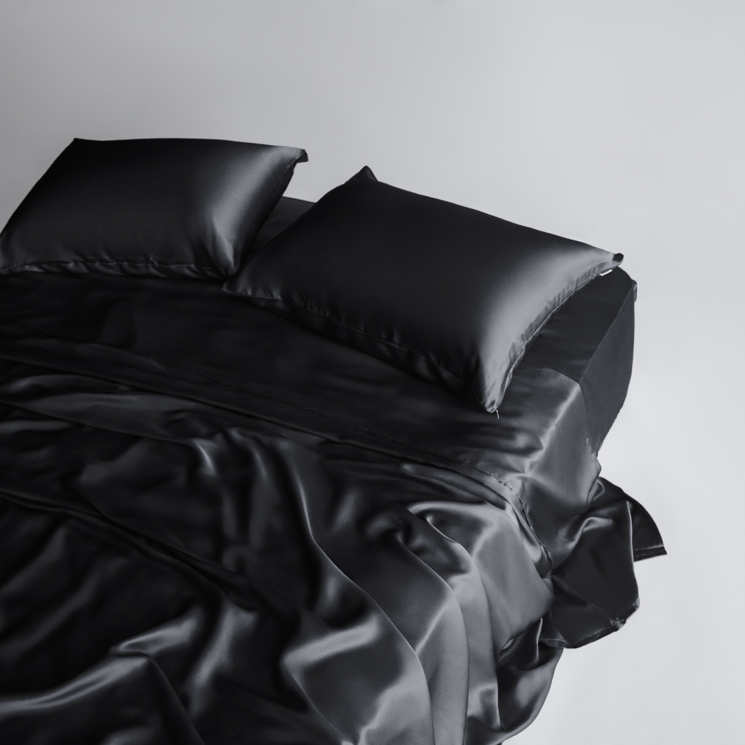 The Complete Sheet Set (4-Piece) features silky soft black sheets and two matching pillows, styled on a neatly made bed against a plain white background for a sleek, modern look.