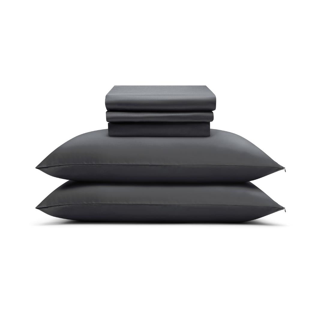 Two neatly stacked charcoal pillows with the Complete Sheet Set (4-Piece) - folded silky soft sheets and pillowcases - arranged on top, all set against a plain white background.
