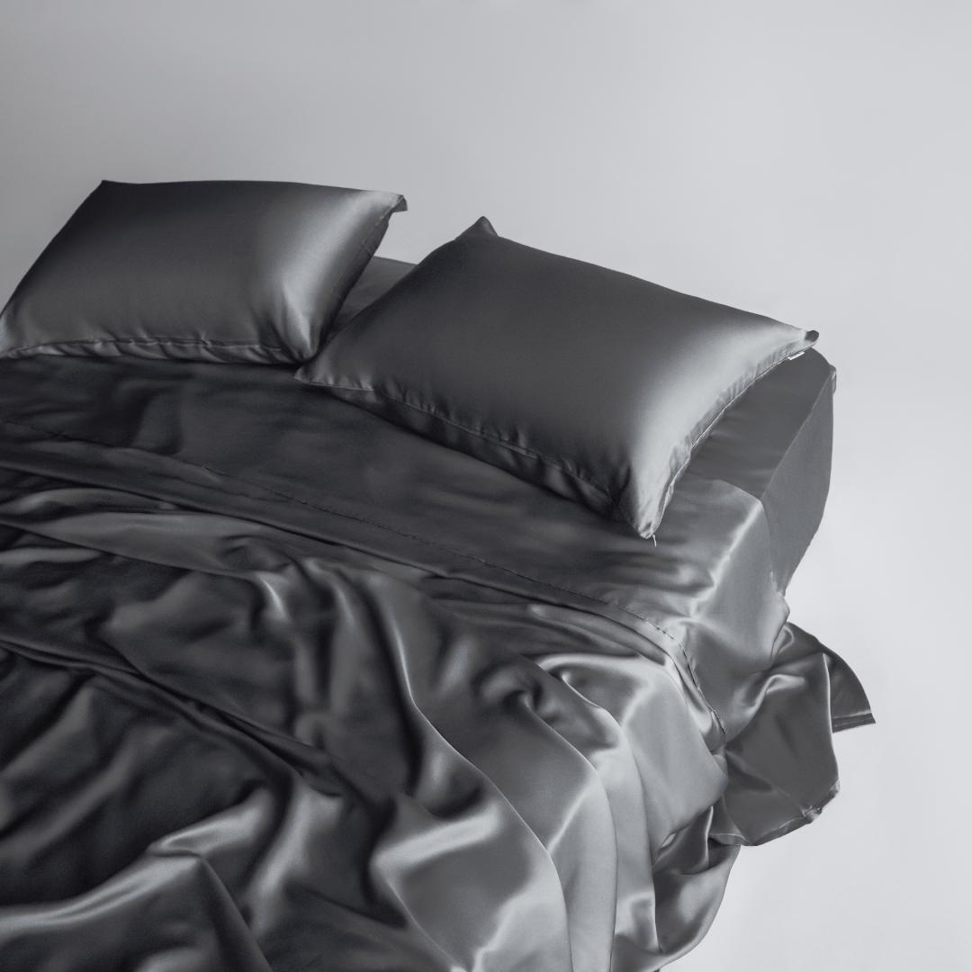 A neatly made bed with two pillows features the Complete Sheet Set (4-Piece) in smooth, silky soft charcoal fabric against a plain light background.
