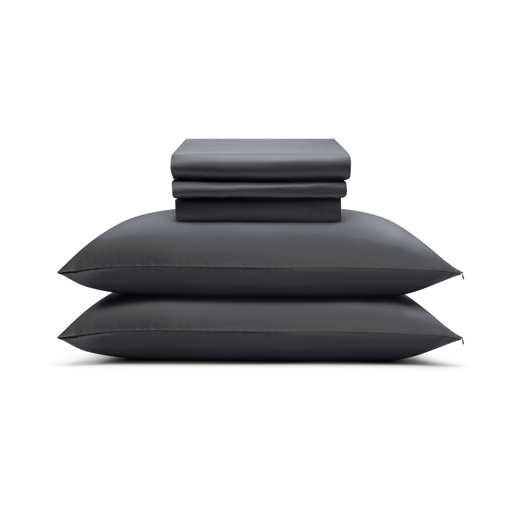 Two neatly stacked black pillows with the Complete Sheet Set (4-Piece)—folded silky soft sheets and pillowcases—arranged on top, all set against a plain white background.