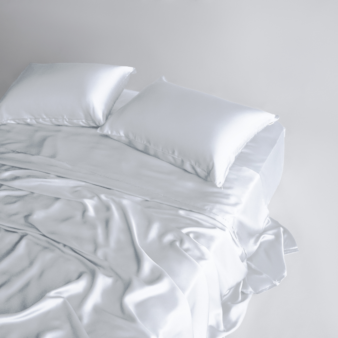 A neatly made bed with two white pillows and slightly rumpled, silky soft Complete Sheet Set (4-Piece) sheets in a bright, minimalistic setting.
