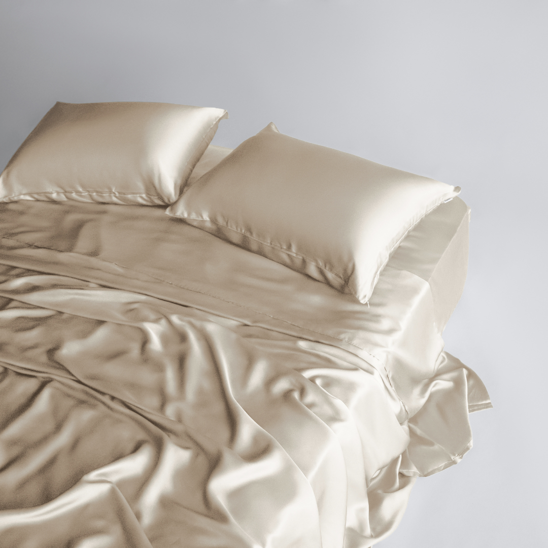 A neatly made bed features the Complete Sheet Set (4-Piece) in light beige satin, with silky soft sheets and two matching satin pillows, set against a plain light background.