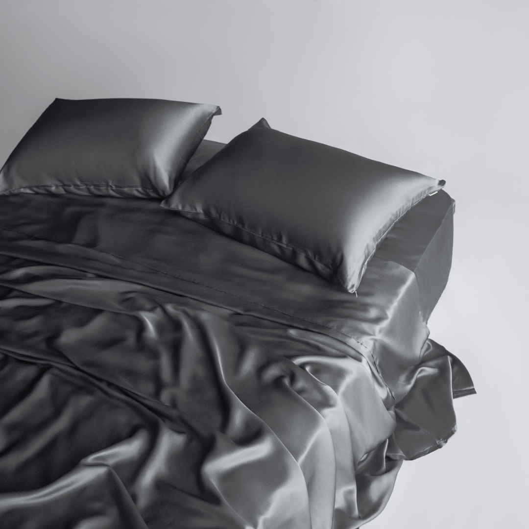 A neatly made bed with two pillows features the Complete Sheet Set (4-Piece) in smooth, silky soft dark gray satin against a plain light background.