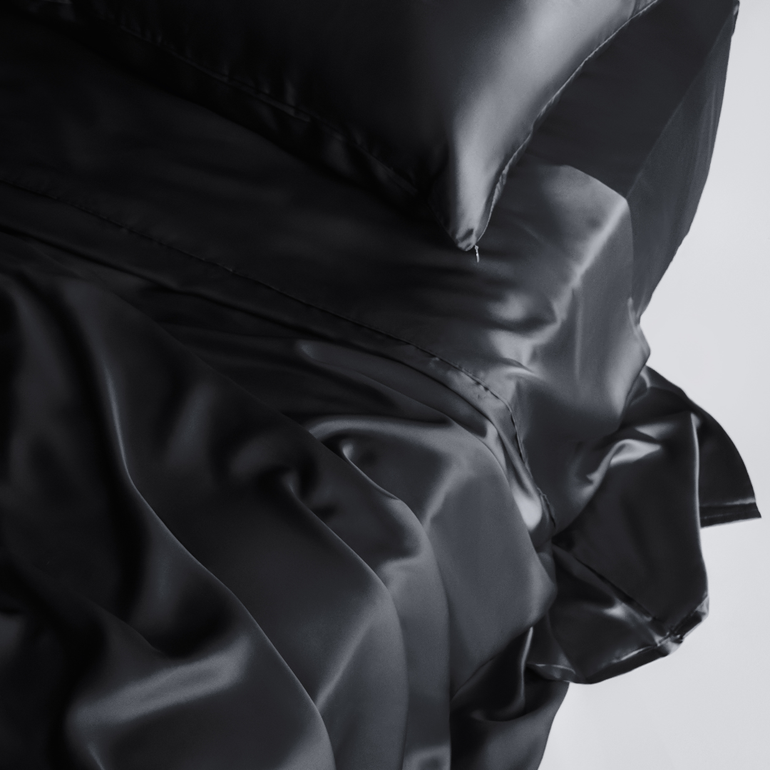 A close-up of a neatly made bed features the Complete Sheet Set (4-Piece) with smooth, silky sheets and glossy black satin pillowcases, showcasing their luxurious texture and elegant shine.