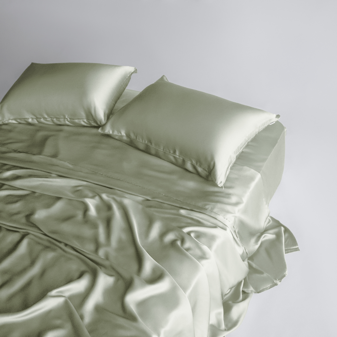A neatly made bed features the Complete Sheet Set (4-Piece) in light sage green, with two pillows and silky satin sheets draped smoothly, adding a touch of luxury to the simple, minimalistic room.