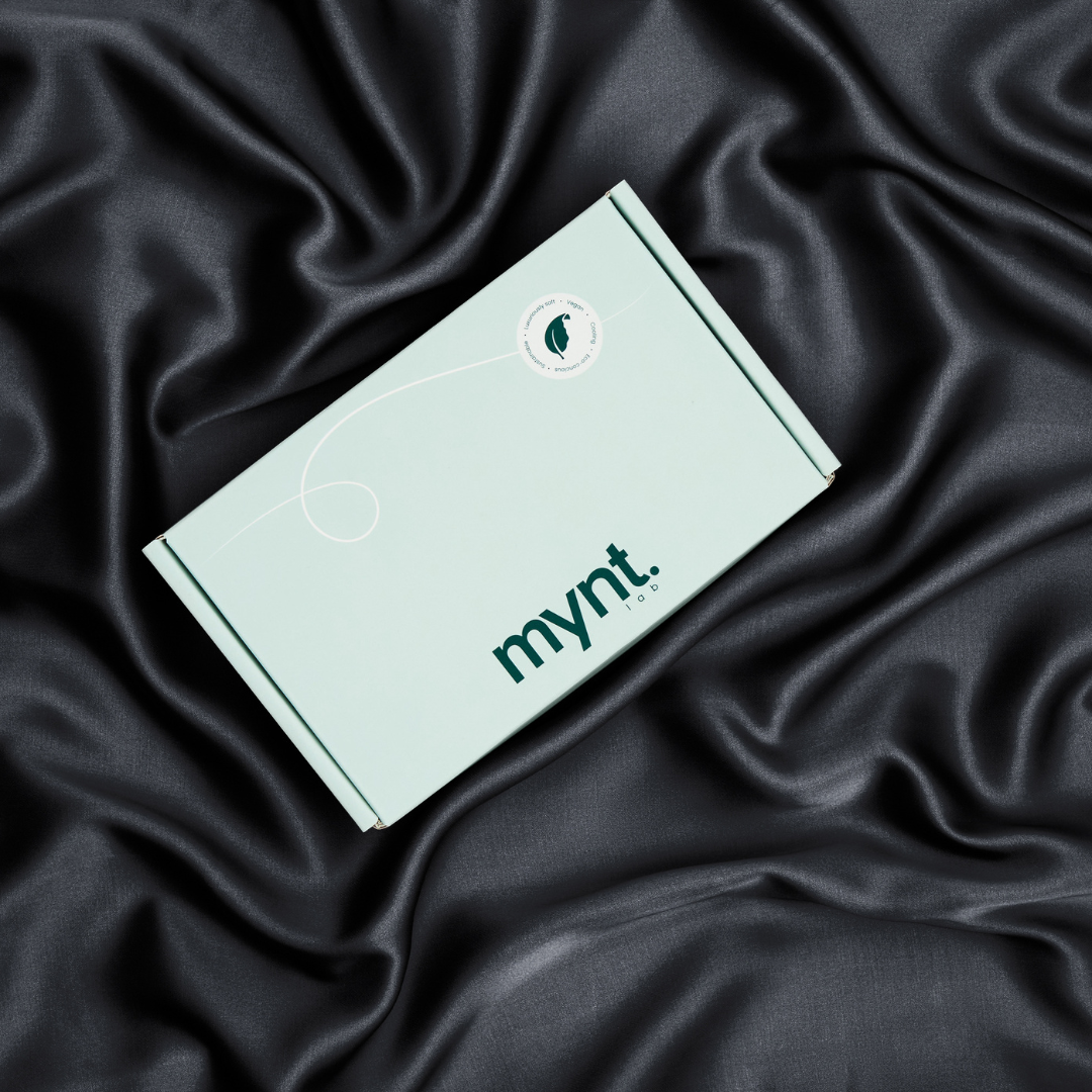 A mint green box with the mynt lab logo rests on black silky fabric. Ideal for a cooling pillowcase or blackout sleep mask,.