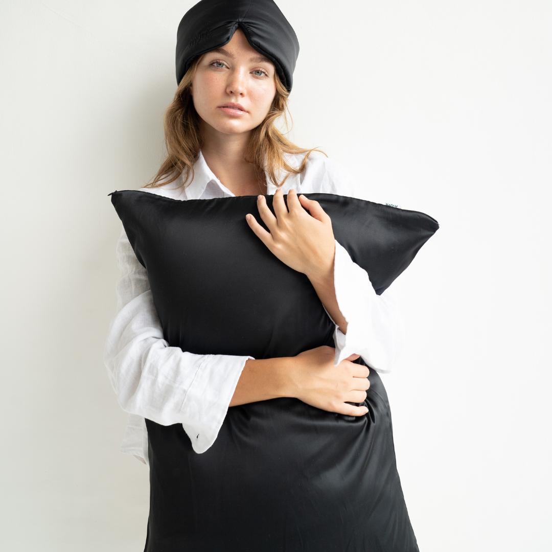 A woman in a black sleep mask and white shirt stands against a white background, holding the Deep Sleep Bundle pillowcase to her chest and looking at the camera with a neutral expression.