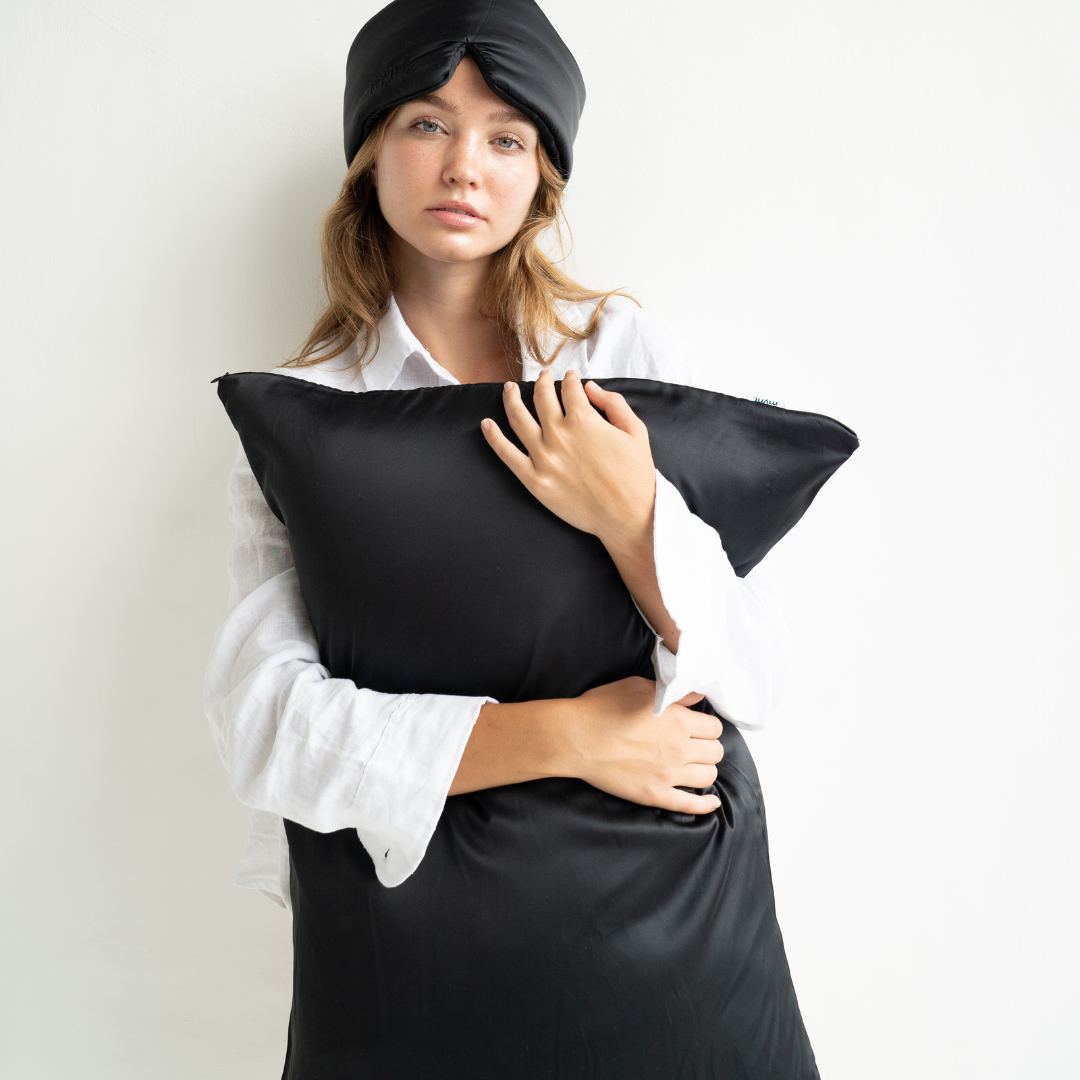 A woman in a black sleep mask and white shirt stands against a white background, holding the Deep Sleep Bundle pillowcase to her chest and looking at the camera with a neutral expression.