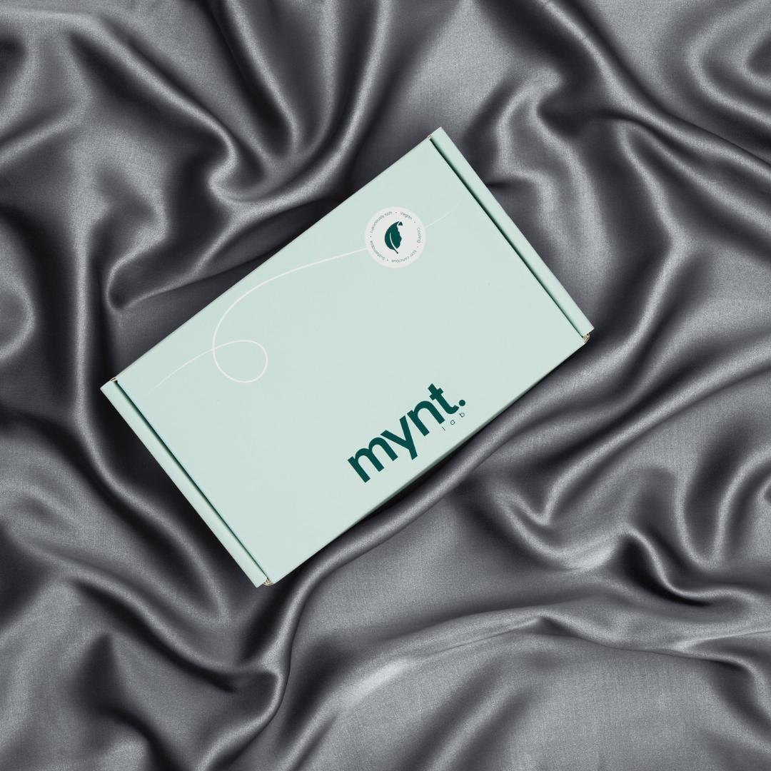 A mint green rectangular box labeled “mynt lab” rests on a smooth, grey silky fabric, contrasting matte and shine. The Deep Sleep Bundle is ideal for anyone wanting restful nights with a cooling pillowcase and blackout sleep mask.
