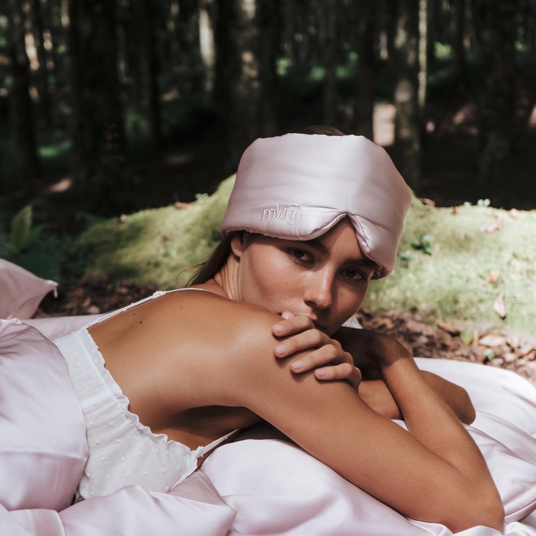 A woman enjoys deep sleep outdoors, wearing the Deep Sleep Bundle’s light pink blackout mask and a white camisole while lying on a silky sheet, surrounded by trees and gentle natural light.