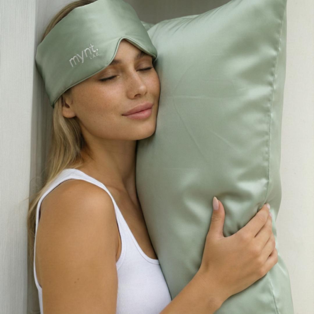 A woman smiles softly, eyes closed, wearing a sage green mask from the Deep Sleep Bundle and hugging its matching silky pillow, dressed in a white tank top and leaning against a white wall.