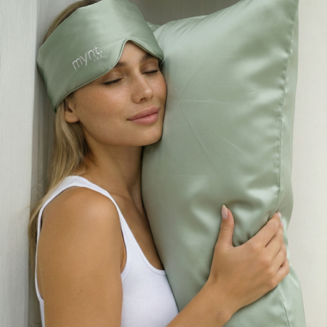 A woman smiles softly, eyes closed, wearing a sage green mask from the Deep Sleep Bundle and hugging its matching silky pillow, dressed in a white tank top and leaning against a white wall.