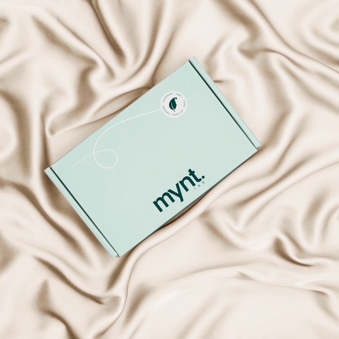 A mint green cardboard box with "mynt lab" printed on it rests on soft beige silky fabric, making the Deep Sleep Bundle - a blackout sleep mask and cooling pillowcase - an ideal gift for enhancing restful sleep.