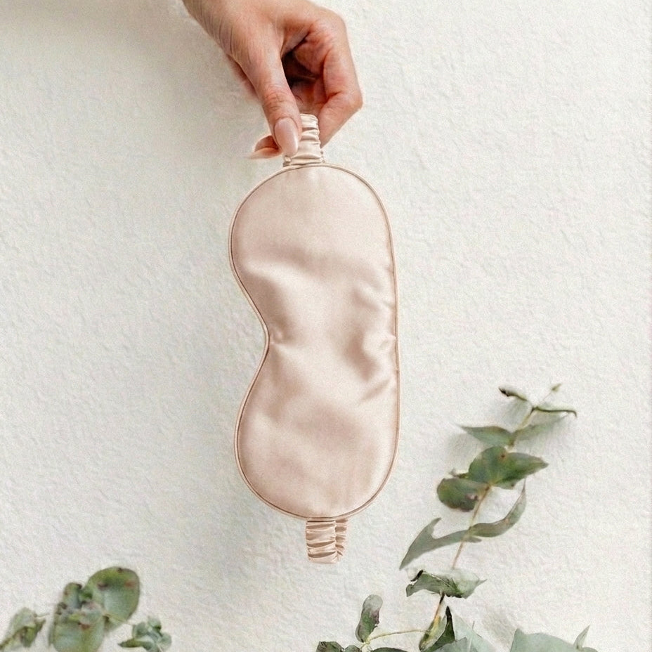 Pink silky eye mask held by a hand with green leaves on a light background