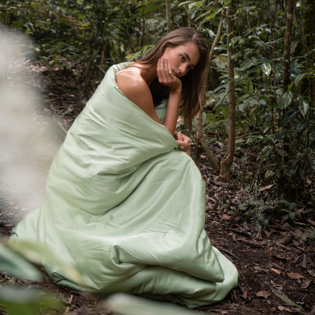 A woman sits on the forest floor wrapped in a light green sustainable Duvet Cover, resting her chin on her hand and gazing thoughtfully at the camera amidst greenery and trees.