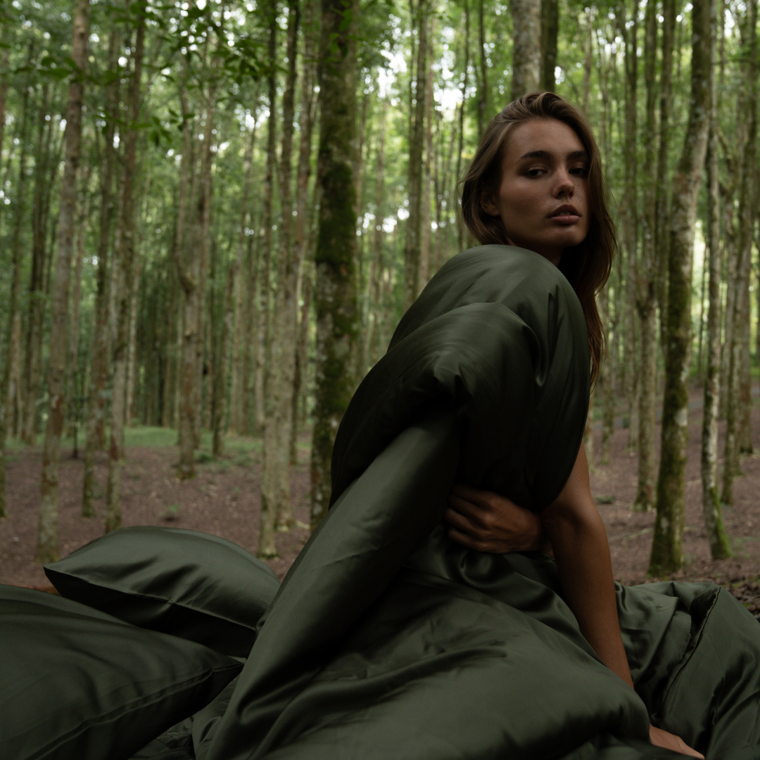 A woman sits outdoors in a forest, wrapped in the Duvet Cover with dark green bedding and green pillows beside her. Surrounded by trees, she looks over her shoulder towards the camera.