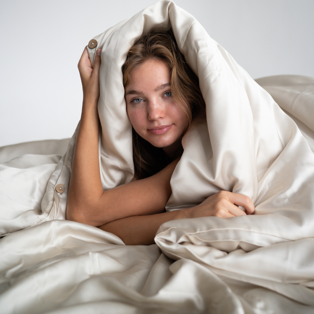 A young woman with long hair smiles softly while wrapped in the Duvet Cover, a silky, cream-colored cooling bedding, as she lies on a bed against a plain white background.