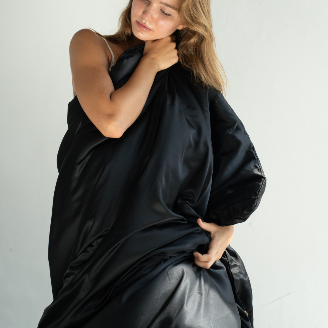 A woman with light brown hair stands against a plain background, wrapped in a large, shiny black Duvet Cover, holding it close to her body with both hands.