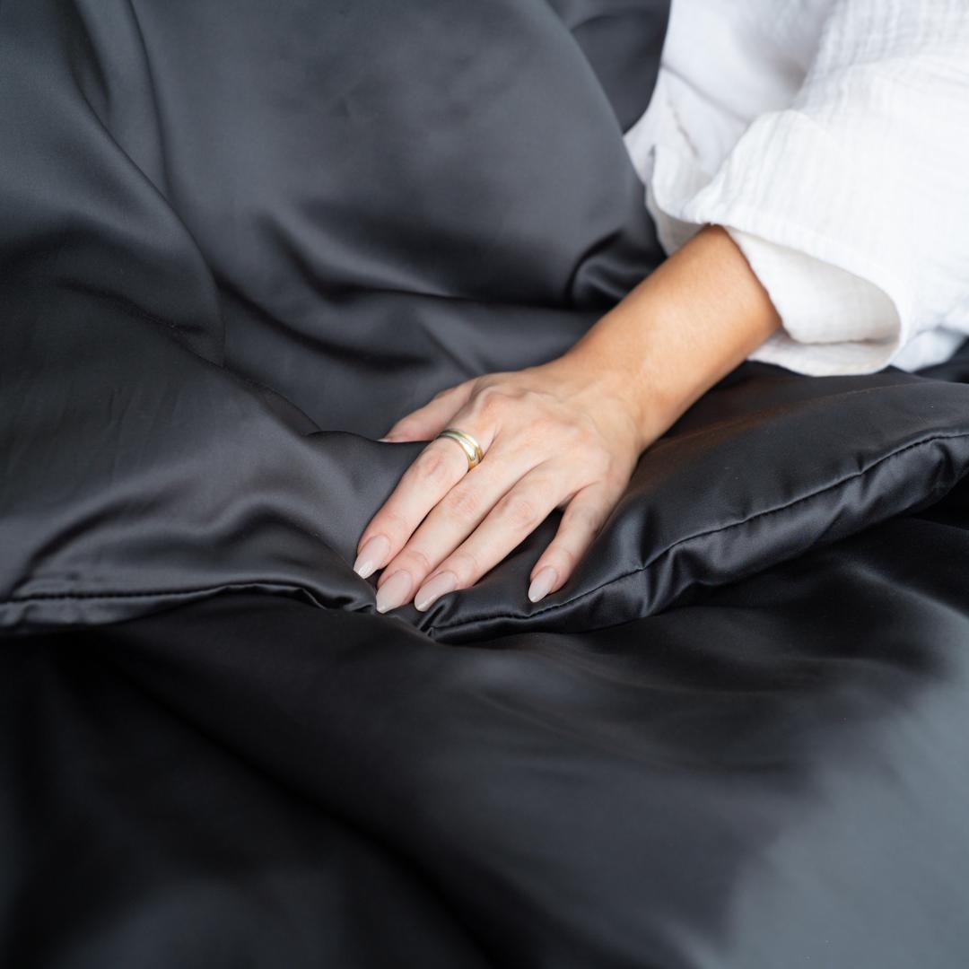 A person with neatly manicured nails and a gold ring rests their hand on a smooth, charcoal-coloured silky Duvet Cover.