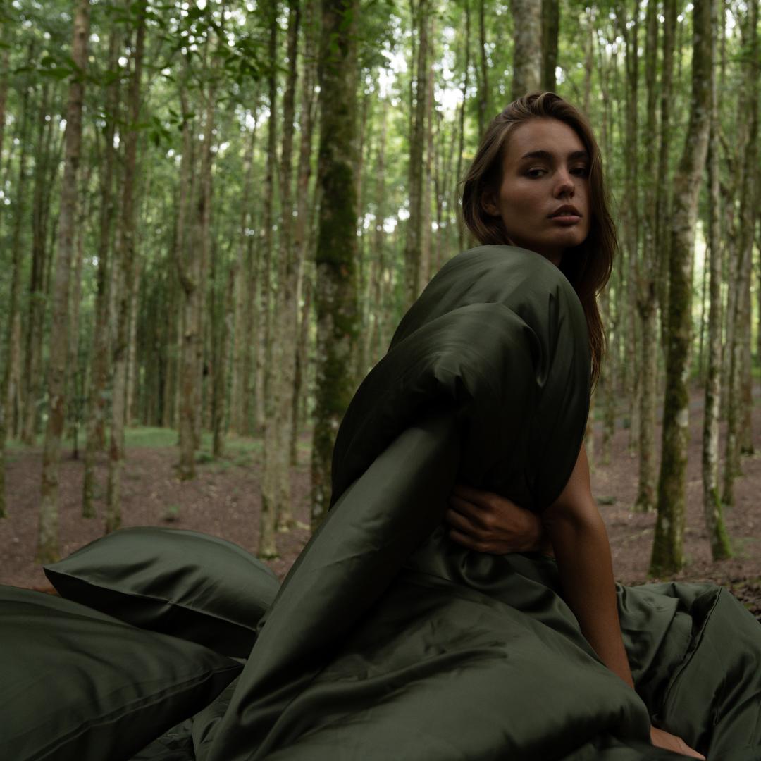 A woman sits outdoors in a forest, wrapped in the Duvet Cover with olive green bedding and pillows beside her. Surrounded by trees, she looks over her shoulder towards the camera.