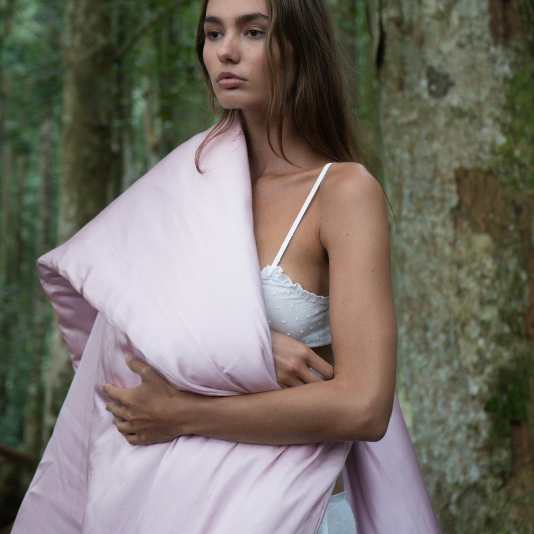 A woman stands in a forest with tall trees, wearing a white polka dot camisole and holding the Duvet Cover, which is sustainable, light pink, and wrapped around her shoulder.