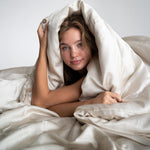 A young woman with long hair smiles softly while wrapped in the Duvet Cover, in silky, sand-coloured cooling bedding, as she lies on a bed against a plain white background.