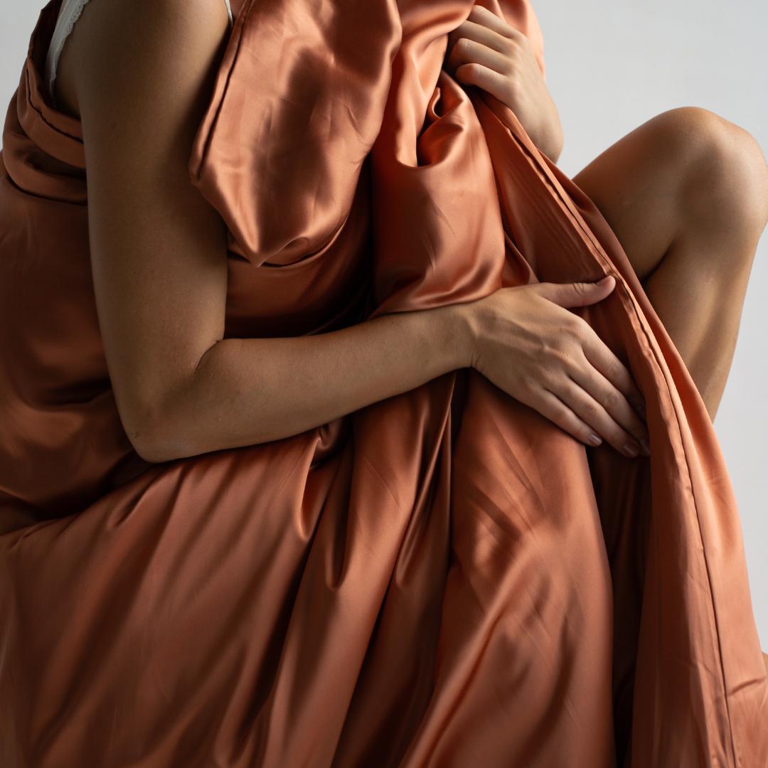 A person draped in a silky, terracotta-coloured fabric sits with one arm on their knee, hand resting on the material - evoking the luxurious comfort of the Duvet Cover. The background is light and neutral.